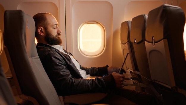 Pasajero masculino que encuentra asiento en el avión para ir de vacaciones, viajando con una aerolínea comercial. Volando en avión durante la puesta de sol, sentado y esperando el aterrizaje en el aeropuerto. Disparo de mano.