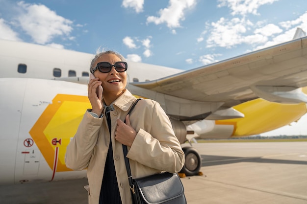 Pasajero sonriente con gafas de sol hablando por teléfono al aire libre en el aeropuerto antes de embarcar