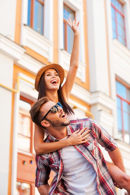 Pasando un buen rato juntos. Hombre joven feliz que lleva a su hermosa novia sobre los hombros y sonriendo mientras camina por la calle