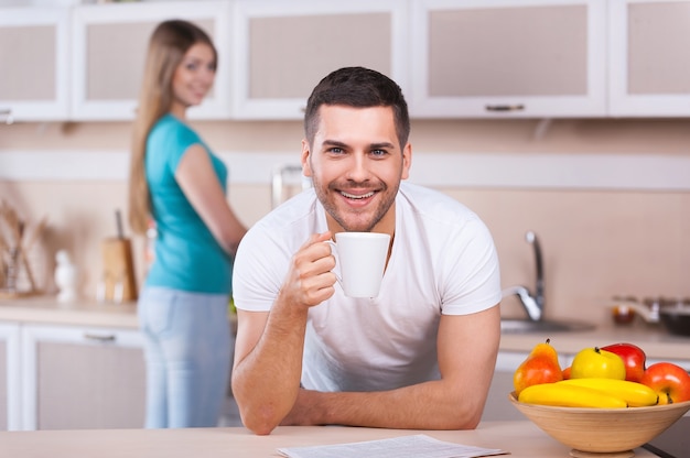 Pasando una buena mañana juntos. Apuesto joven apoyado en la estufa de la cocina y sosteniendo una taza de café mientras hermosa mujer de pie en el fondo y sonriendo