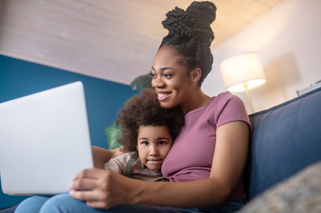 Pasando el tiempo. Mamá afroamericana abrazada y niña linda rizada mirando portátil en casa en el sofá de buen humor