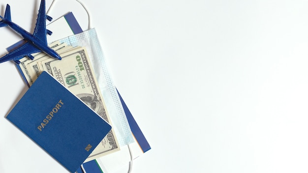 Pasaporte y dinero en efectivo en dólares, máscara y estatuilla de avión sobre un fondo blanco.
