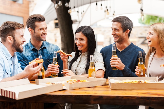 Pasar un buen rato con amigos. Grupo de jóvenes alegres comiendo pizza y bebiendo cerveza mientras está de pie al aire libre