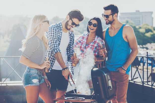 Pasar un buen rato con amigos. Grupo de jóvenes alegres que se unen entre sí y hacen una barbacoa mientras están de pie en el techo