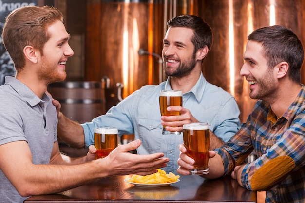 Pasar un buen rato con amigos. Tres hombres jóvenes felices en ropa casual hablando y bebiendo cerveza mientras están sentados juntos en el pub de cerveza