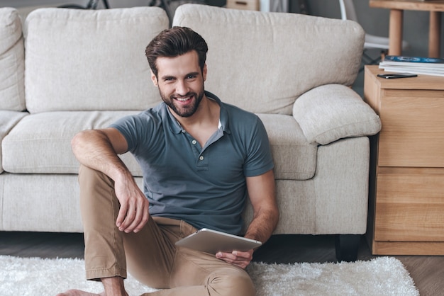 Pasar un buen rato en casa. Apuesto joven sosteniendo su tableta digital y mirando a la cámara con una sonrisa mientras está sentado en la alfombra en casa