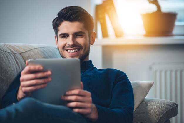 Pasar un buen rato en casa. Primer plano de un joven apuesto con su tableta digital con una sonrisa