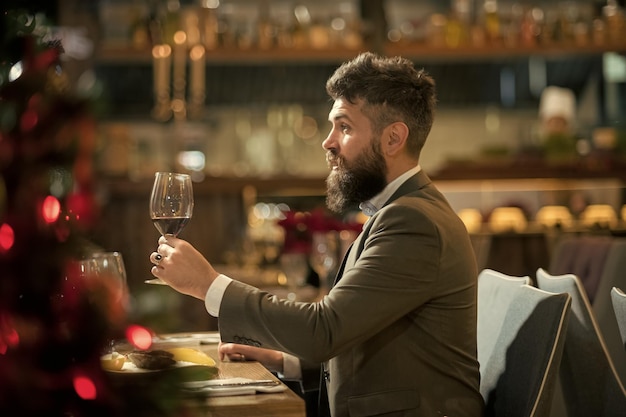 Pasar un buen rato en el restaurante Apuesto joven sosteniendo un vaso con vino tinto y sonriendo en el restaurante