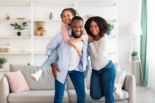 Pasar tiempo con la familia es divertido. Retrato de feliz negro hombre, mujer y niña posando a la cámara, de pie en la sala de estar en casa. Padre alegre que lleva a su hija en la espalda, dando caballito