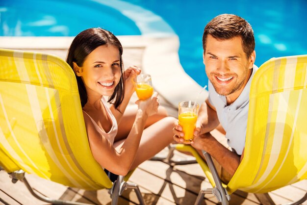 Pasar tiempo sin preocupaciones junto a la piscina. Feliz pareja sosteniendo cócteles y sonriendo mientras está sentado en las tumbonas junto a la piscina