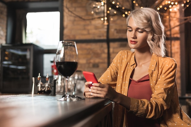 Pasatiempo agradable. Encantadora mujer joven sentada en la barra del bar y navegar por Internet mientras bebe una copa de vino tinto