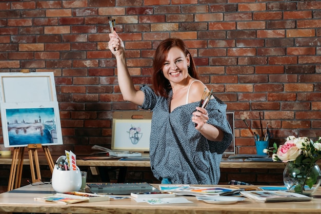 Foto pasatiempo artístico. ambiente de espacio de trabajo de estudio. mujer pelirroja entusiasta posando con pinceles y caballete