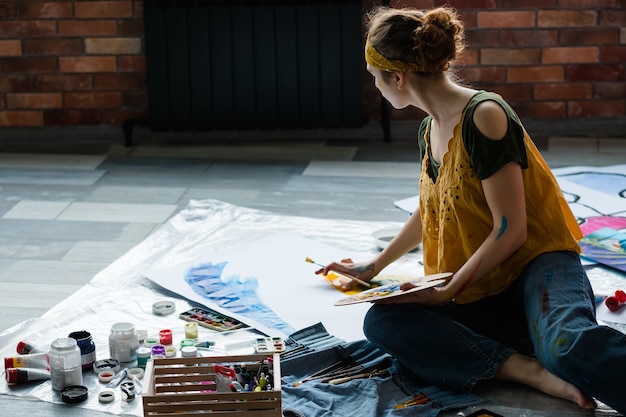 Foto pasatiempo artístico y recreación. artista femenina sentada en el suelo, con paleta de pintura acrílica, creando obras de arte abstractas.