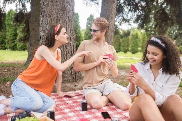 Foto pasatiempo, juego. chico pelirrojo alegre joven y dos niñas sonrientes de pelo largo jugando a las cartas en la naturaleza en buen día