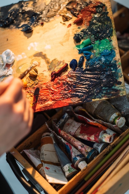 Pasatiempo de pintura Proceso de arte Suministros de pintor Artista femenina mezclando a mano colores de pintura acrílica con pincel en una vieja paleta de madera sucia en un lugar de trabajo desordenado con tubos