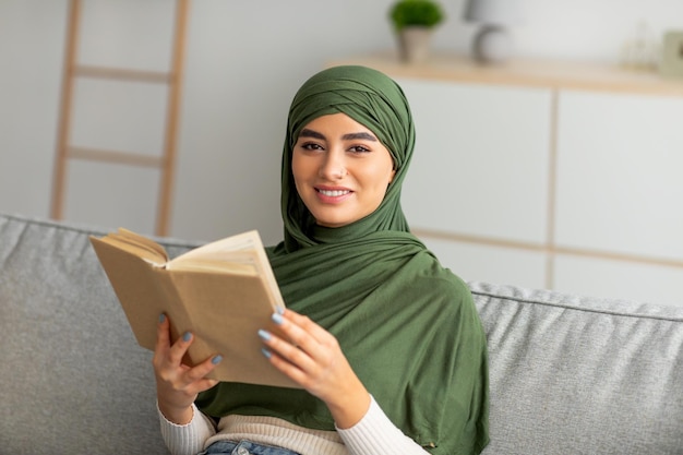 Pasatiempos acogedores en casa Joven mujer árabe con hiyab leyendo un libro sentada en un sofá en la sala de estar