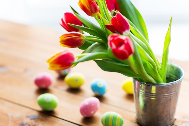 páscoa, feriados, tradição e conceito de objeto - close-up de ovos de páscoa coloridos e flores de tulipa no balde na mesa de madeira