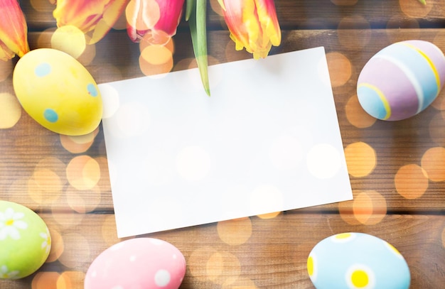 páscoa, feriados, tradição e conceito de objeto - close-up de ovos de páscoa coloridos, flores de tulipa e cartão de papel branco em branco na superfície de madeira com espaço de cópia sobre luzes de férias