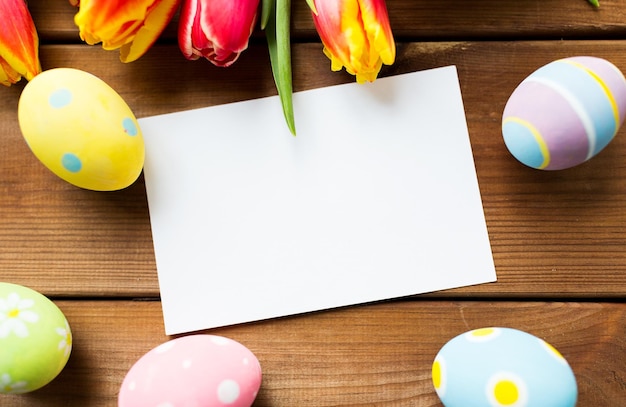páscoa, feriados, tradição e conceito de objeto - close-up de ovos de páscoa coloridos, flores de tulipa e cartão de papel branco em branco na superfície de madeira com espaço de cópia