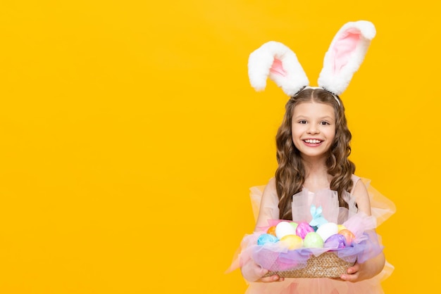 Páscoa festiva para crianças Uma linda adolescente com cabelos cacheados segura uma cesta de ovos coloridos para o feriado Uma criança fantasiada de coelho com orelhas compridas em um fundo amarelo isolado