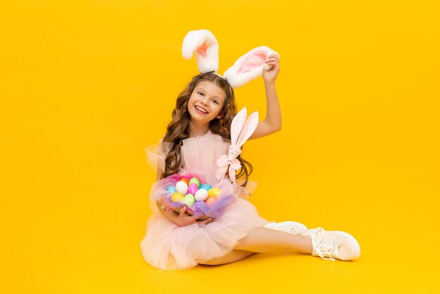 Páscoa festiva Uma garotinha com orelhas de coelho e uma cesta de ovos coloridos sorri amplamente em um fundo amarelo isolado