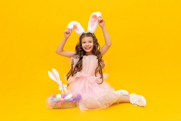 Páscoa festiva Uma menina de cabelos cacheados com orelhas de coelho em um vestido rosa está aproveitando o feriado Uma criança com uma cesta de ovos coloridos em um fundo amarelo isolado