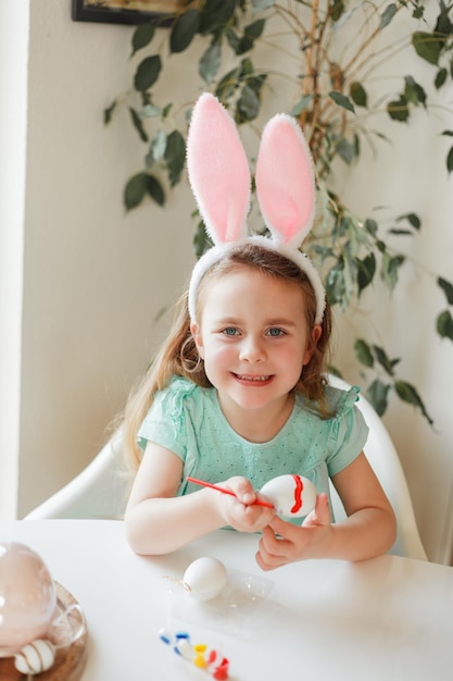 Páscoa Menina bonitinha em orelhas de coelho decora ovos de Páscoa na mesa em casa nas férias de primavera
