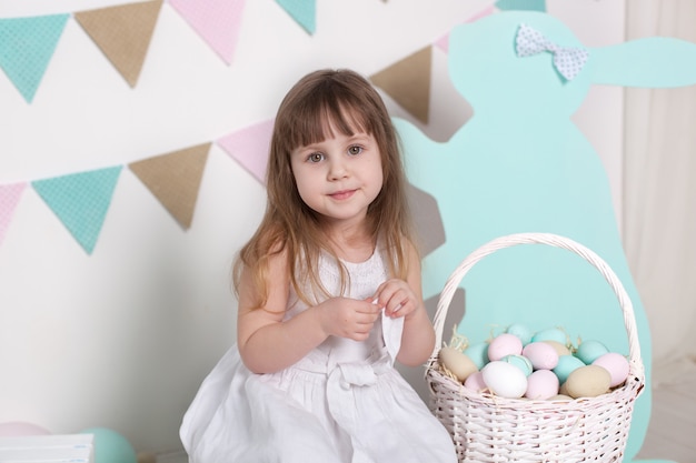 Páscoa. menina de vestido branco está sentado perto da cesta com ovos e um coelhinho da páscoa. localização de páscoa colorida, decoração de primavera. férias em família, tradições. fazendeiro. colheita. celebração
