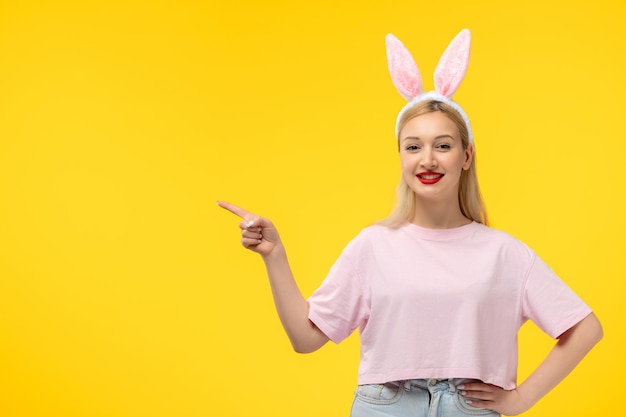 Pascua adorable jovencita rubia con orejas de conejo sonriendo y apuntando hacia arriba