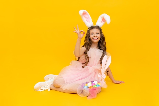 Pascua festiva Una niña feliz está sentada y disfrutando de las vacaciones de primavera Un niño con orejas de conejo sobre un fondo amarillo aislado