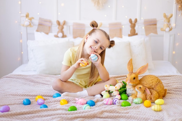 Pascua, una niña pequeña con huevos de colores pintados, tulipanes y un conejo en casa en una habitación luminosa se prepara para las vacaciones sonriendo, divirtiéndose y jugando con huevos cerrando los ojos