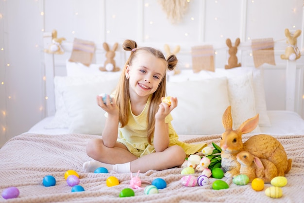 Pascua, una niña pequeña con huevos de colores pintados, tulipanes y un conejo en casa en una habitación luminosa se prepara para las vacaciones sonriendo, divirtiéndose y jugando con huevos cerrando los ojos