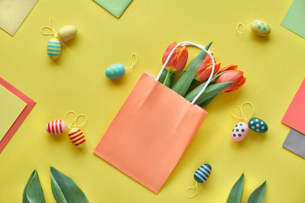 Pascua plana yacía sobre papel amarillo. Ramo de tulipanes, cajas de regalo, huevos decorativos y bolsas de papel, disposición geométrica.
