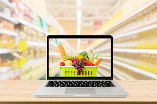 Foto pasillo de supermercado fondo borroso con computadora portátil y carrito de compras en el concepto de línea de comestibles de mesa de madera