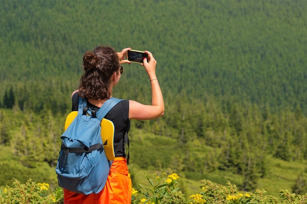La pasión por los viajes de la mujer que fotografía el paisaje de la naturaleza del paisaje en smartphone en el bosque. Hembra joven que toma la foto en celular. Fotógrafo de naturaleza turista dispara mientras está parado en la cima de la montaña.