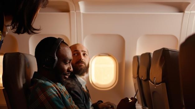 Passageiro masculino conversando com comissário de bordo no avião, sentado no avião e usando smartphone. Voando durante o pôr do sol, viajando em viagem de férias com turistas.