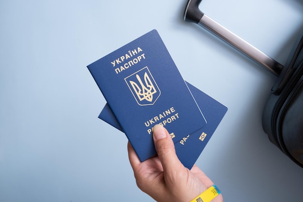 Passaportes de um cidadão da Ucrânia em uma mão feminina em um fundo azul com mala closeup Conceito de emigração Inscrição no passaporte ucraniano da Ucrânia