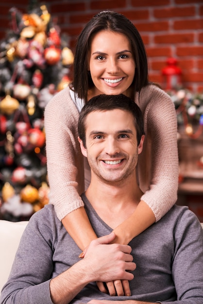 Passar o Natal juntos. Belo jovem casal apaixonado, unindo-se e sorrindo com uma árvore de Natal ao fundo