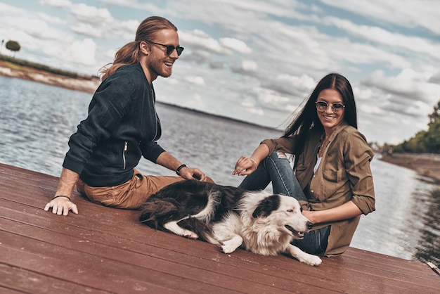 Passar tempo juntos. Lindo casal jovem brincando com um cachorro sentado perto do lago ao ar livre