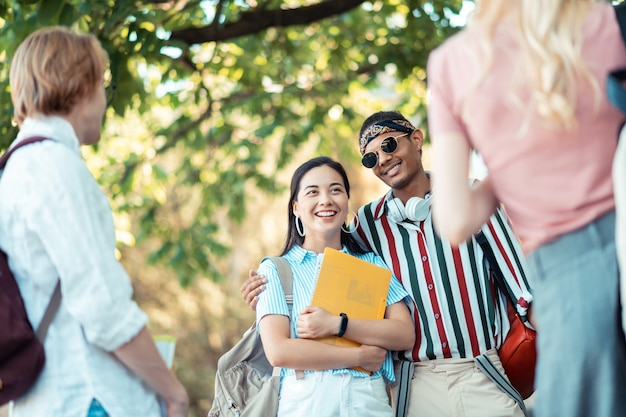 Passar tempo juntos. rapaz alegre, vestindo roupas brilhantes, abraçando sua namorada esperta com livros nas mãos e conversando com seus amigos.