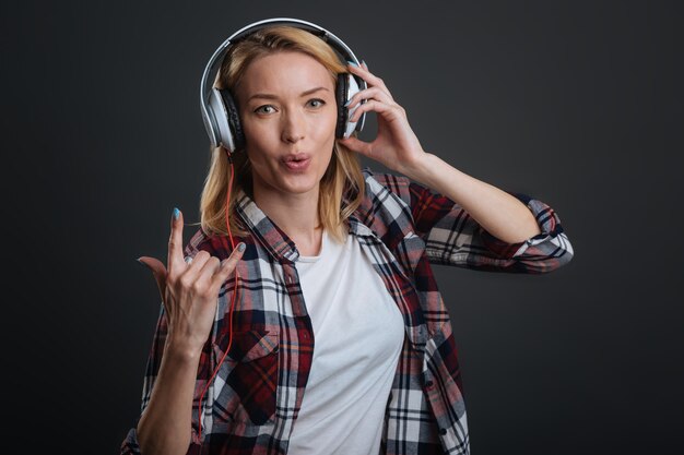 Passatempo agradável. Jovem graciosa bonita usando fones de ouvido e ouvindo música enquanto fica de pé, isolado em um fundo cinza