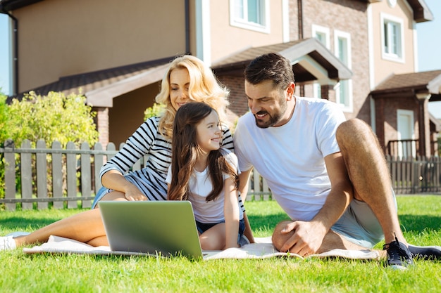 Passatempo agradável. Jovens pais amorosos e sua filha sentados no tapete no quintal perto de sua casa e assistindo desenhos animados juntos no laptop