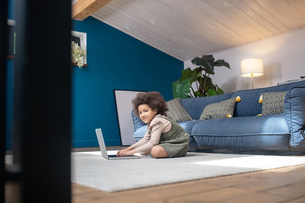 Passatempo, laptop. garotinha sorridente de pele escura sentada com o laptop no chão em um quarto moderno e aconchegante em casa