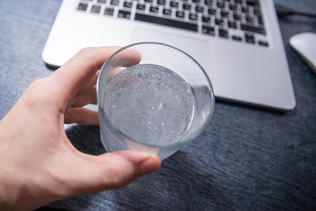 Foto passe o comprimido de aspirina em um copo d'água. laptop em segundo plano. sobrecarregado com trabalho de escritório