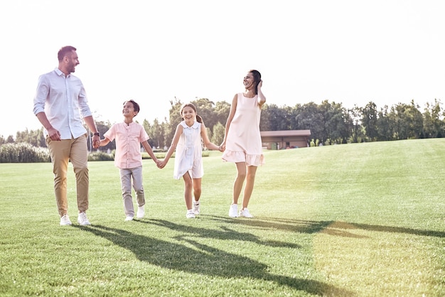 Passeio em família. Família de quatro pessoas caminhando no campo gramado, olhando um para o outro e rindo de felicidade