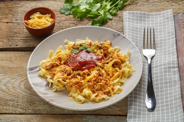 Pasta Bolognese garniert mit Kräutern und Käse in einem Teller auf einem Holztisch neben einer Gabel und einer Serviette und Gemüse auf einem Brett und geriebenem Käse