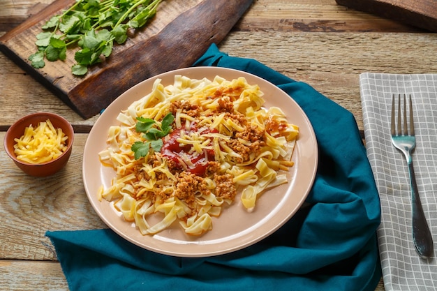 Pasta Bolognese garniert mit Kräutern und Käse in einem Teller auf einer blauen Serviette auf einem Holztisch neben einer Gabel und Gemüse auf einem Brett