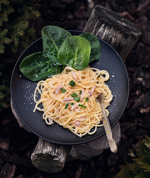 Pasta carbonara decorada con hojas de espinacas y parmesano en la placa negra. Cena tradicional en cursiva en el jardín rústico.