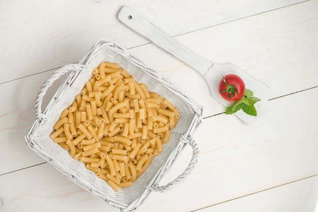 Pasta en cesta de mimbre y tomate en una cuchara de madera sobre una vieja tabla de madera blanca