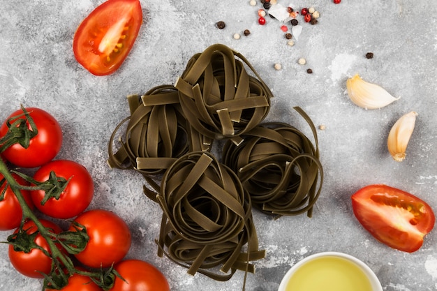 Pasta cruda de tallarines con espinacas e ingredientes para cocinar (tomates cherry, especias, ajo) sobre fondo gris. Vista superior. Fondo de comida